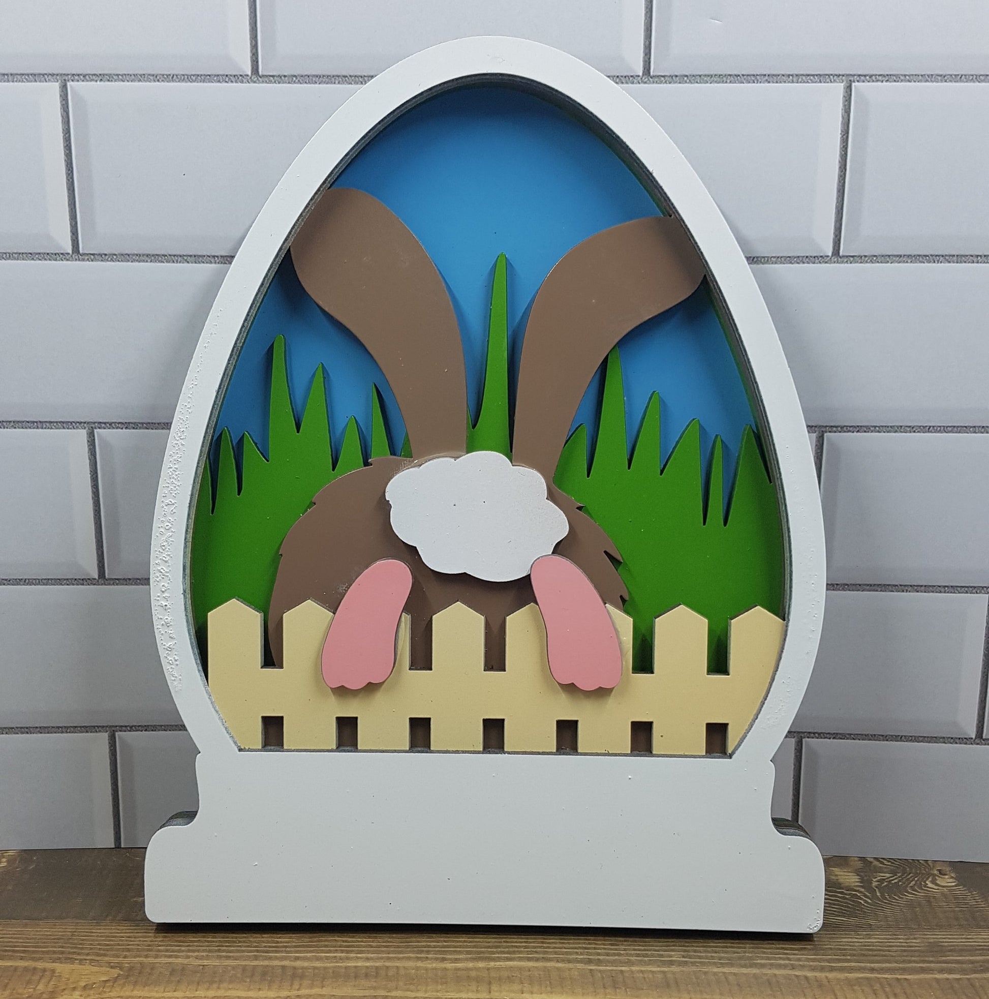 Explore our Easter-themed 5-layered CO2 laser-cut sign: Large (15.7cm x 20cm). Adorable bunny hops over rustic fence into serene garden scene against blue sky backdrop. Meticulously crafted from high-quality materials. Perfect for Easter decor enthusiasts. Celebrate Easter joy with Turtle and Wolf Makers and Crafters.