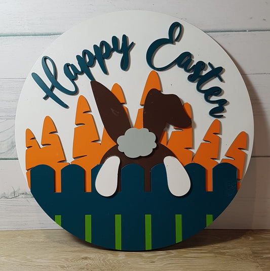 round Happy Easter sign with a white background showing a brown Easter Bunny's ears, white feet and grey tail hopping over a garden fence into a carrot patch. The sign is laser cut, with 4 layers for that added dimensional effect. Dimensions 30cm x 30cm.