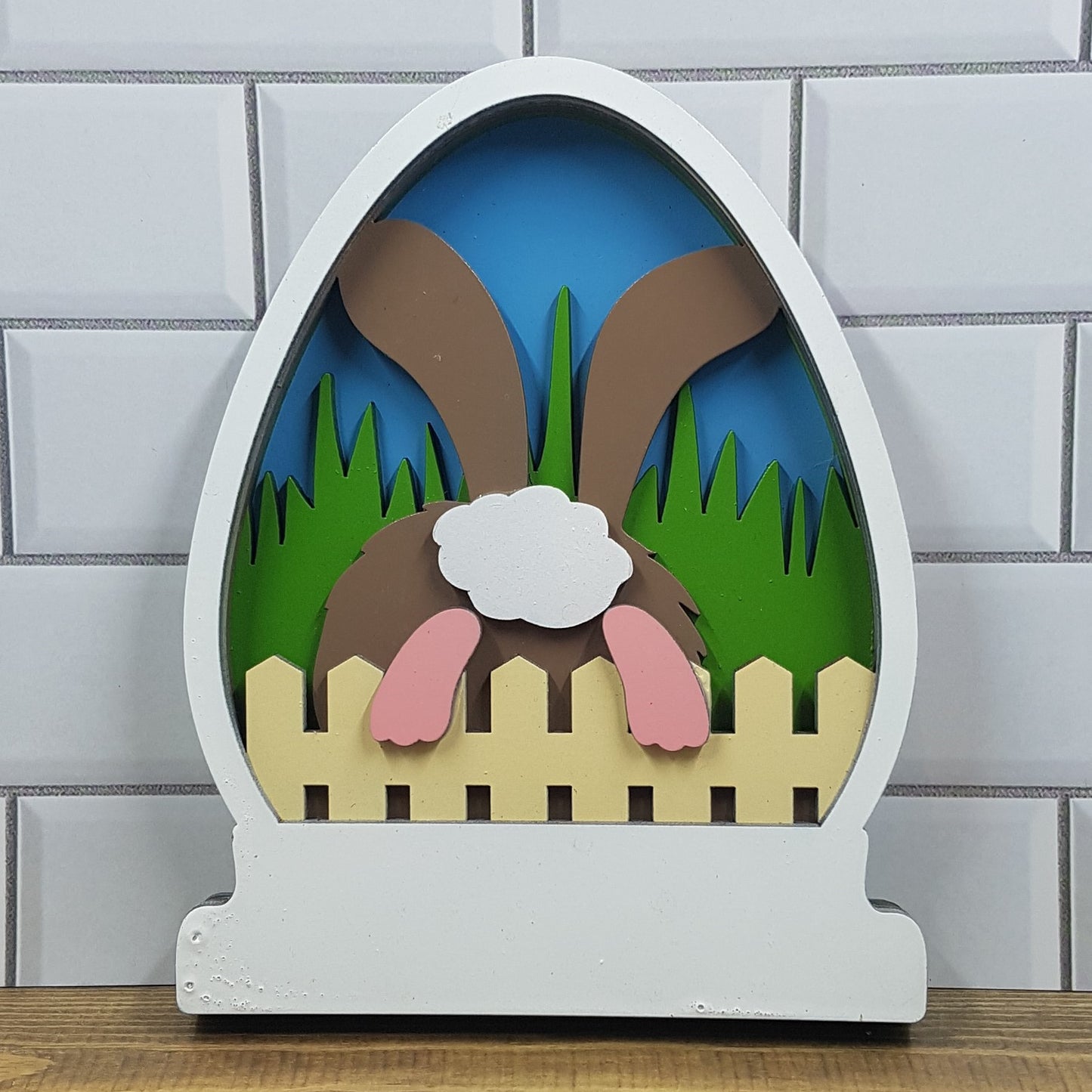 Discover our Easter-themed 5-layered CO2 laser-cut sign: Small (12cm x 15cm). Adorable bunny hops over rustic fence into serene garden scene against blue sky backdrop. Meticulously crafted from high-quality materials. Perfect for Easter decor enthusiasts. Celebrate Easter joy with Turtle and Wolf Makers and Crafters.